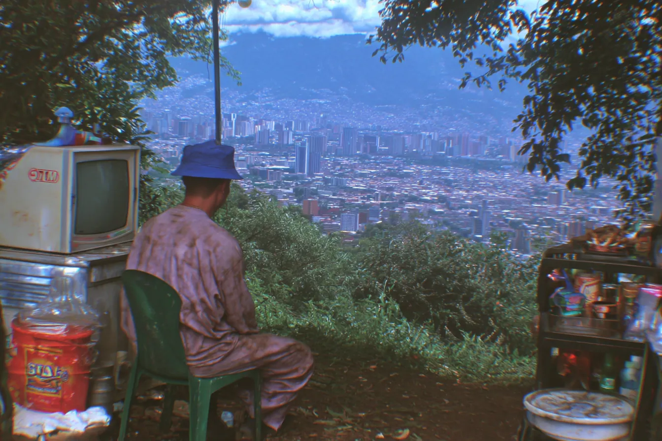 Fotografía de un hombre visto de espaldas, sentado en un banco en una ladera con un televisor y objetos domésticos cerca, mirando una ciudad extensa al fondo.