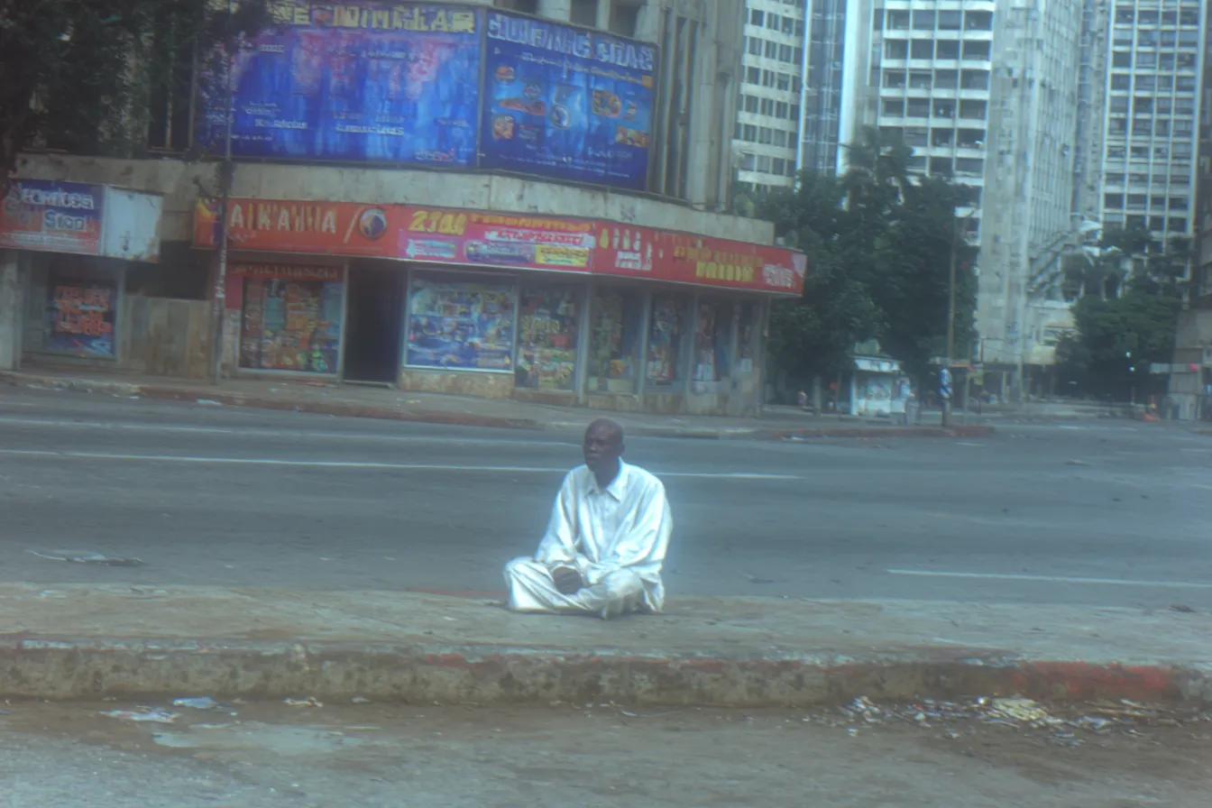 Fotografía de un hombre vestido de blanco sentado con las piernas cruzadas en el separador de una avenida ancha, con tiendas y edificios al fondo.
