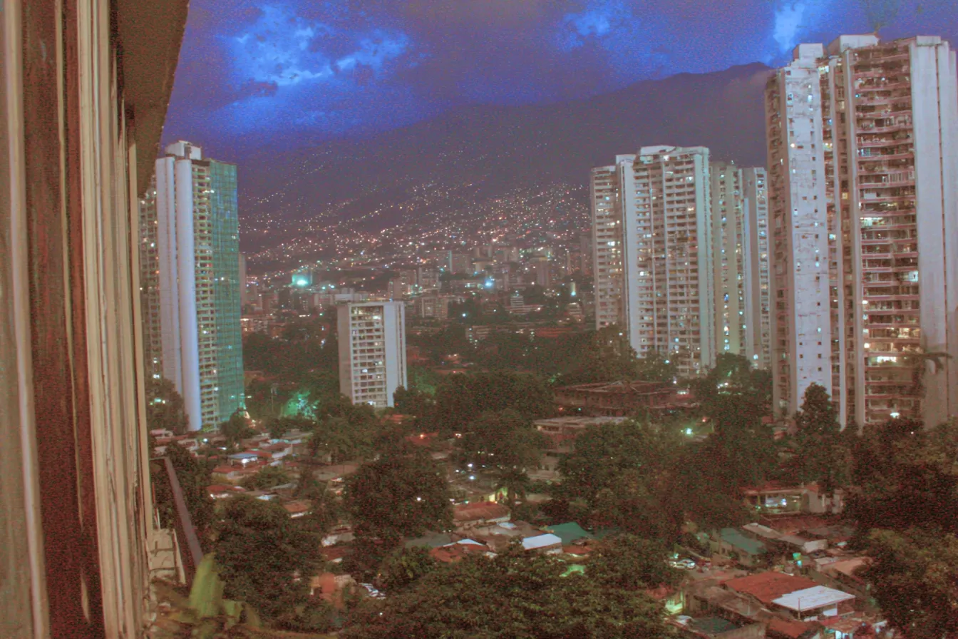 Vista nocturna desde un balcón de una ciudad con edificios altos e iluminados, un cielo azul oscuro cargado de nubes de tormenta, y un barrio de casas humildes con luces cálidas en primer plano.