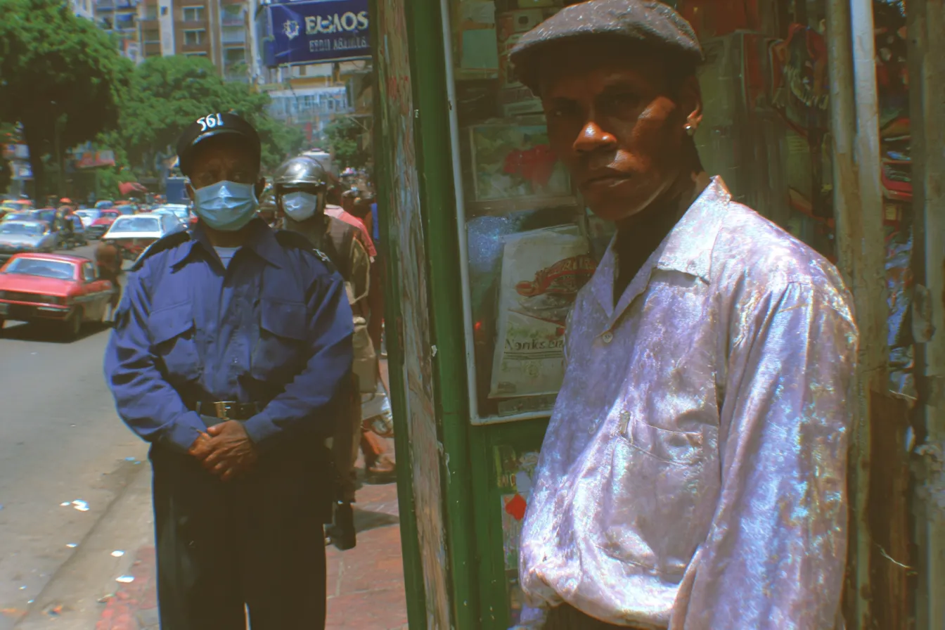 Un hombre con camisa clara salpicada de manchas mira a la cámara junto a un kiosco de madera verde con afiches; detrás, un policía con tapabocas y otra persona con mascarilla caminan por la calle.