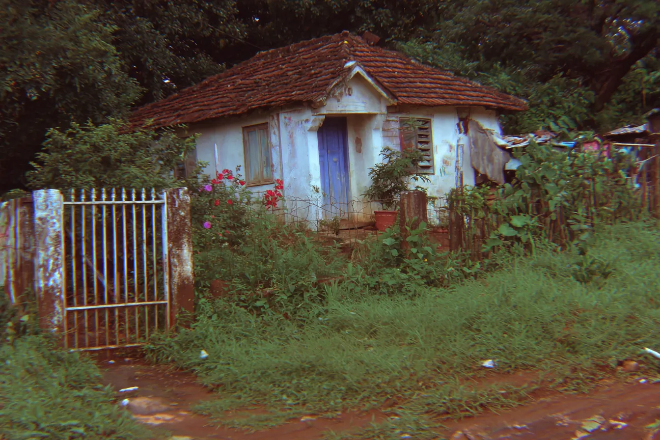 Una casa pequeña con techo de tejas rojas y paredes rosadas descascaradas, puerta y ventanas azules, rodeada de un jardín con maleza y flores rosadas, con un portón de metal oxidado al frente.