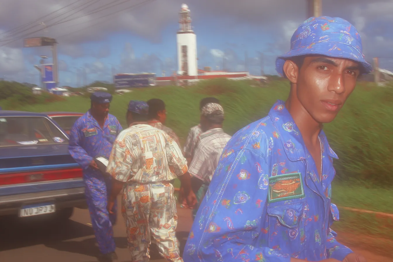 Un hombre con sombrero y camisa de estampado floral azul mira hacia la cámara en primer plano; detrás, varias personas con ropa de camuflaje están junto a un carro, con un faro blanco y un cielo nublado al fondo.