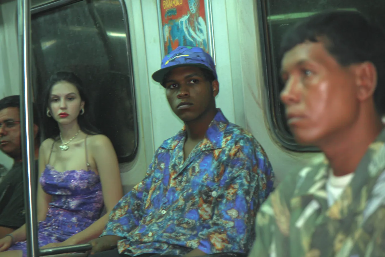 Dentro de un vagón del metro, una mujer con vestido morado y un hombre con camisa de flores azules están sentados uno junto al otro; en primer plano se ve borroso otro pasajero.