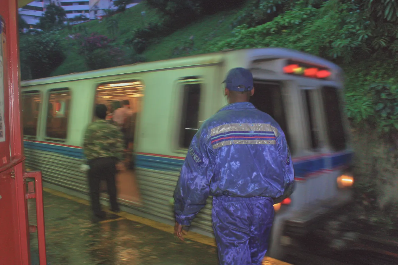 Un hombre con chaqueta y pantalón azul camina hacia un tren del metro que llega a la estación, sobre un andén mojado por la lluvia que refleja las luces; hay una caseta roja a un lado y vegetación en la ladera de arriba.