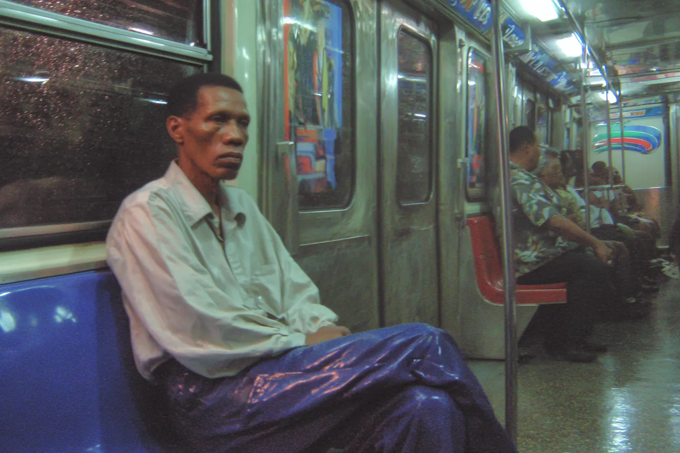 Un hombre con camisa clara y pantalón morado brillante está sentado solo en un asiento del metro; al fondo, otros pasajeros con camisas de colores están sentados en la banca de enfrente.