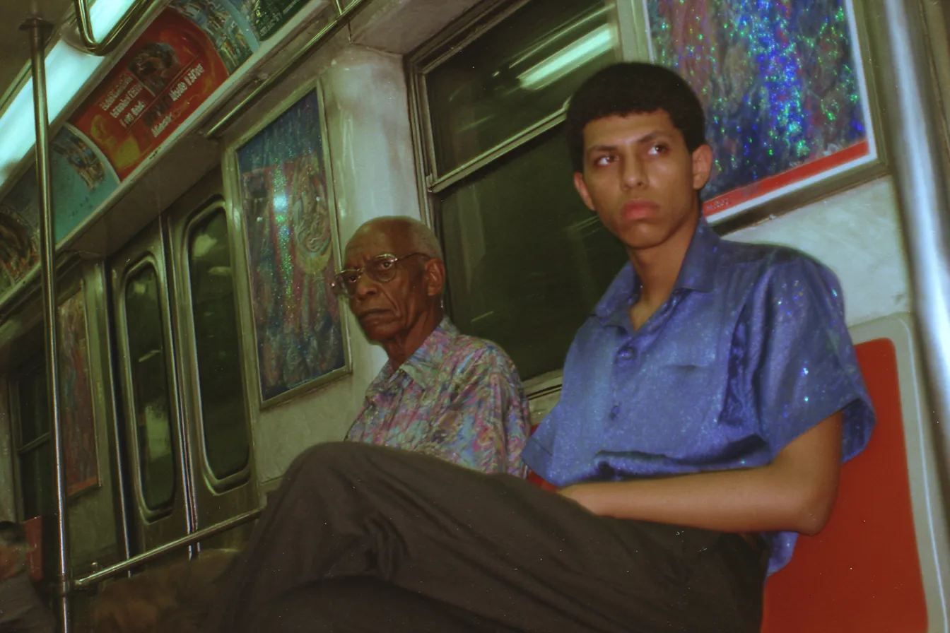 Un hombre mayor con camisa estampada y un joven con camisa azul están sentados uno al lado del otro en un vagón del metro, con afiches de colores en la pared detrás.