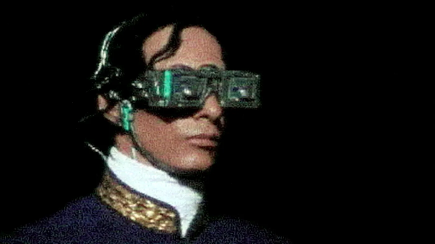 Retrato de una persona con el cabello oscuro peinado hacia atrás, que lleva unos anteojos hechos de placas de circuito electrónico verde sobre los ojos, cuello blanco alto y chaqueta oscura con adornos dorados, sobre fondo negro.