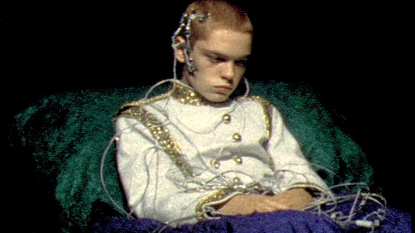 Una persona joven de cabello claro está recostada con los ojos cerrados sobre un cojín verde y una manta morada, con varios cables conectados a la cabeza y vistiendo una chaqueta blanca con adornos dorados.