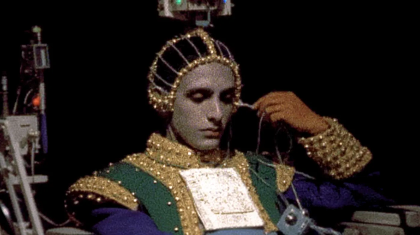 Una persona con los ojos cerrados lleva una corona bordada con perlas y una túnica verde y dorada muy adornada, con cables conectados a la cabeza, mientras sostiene un libro de tapa enjoyada con una mano.