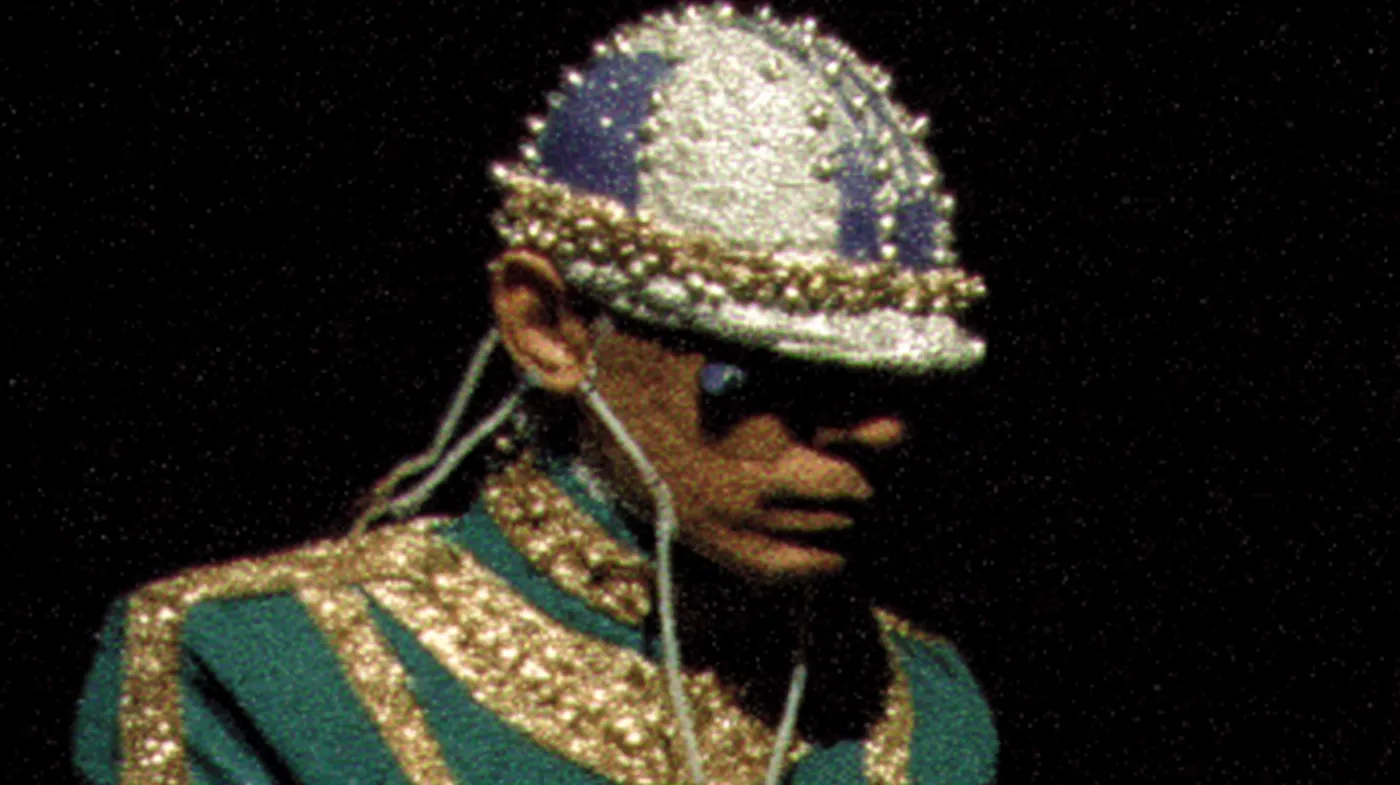 Una persona con gafas oscuras lleva un casco cubierto de piezas brillantes y una chaqueta verde y dorada con bordado de perlas, vista de perfil sobre un fondo oscuro.