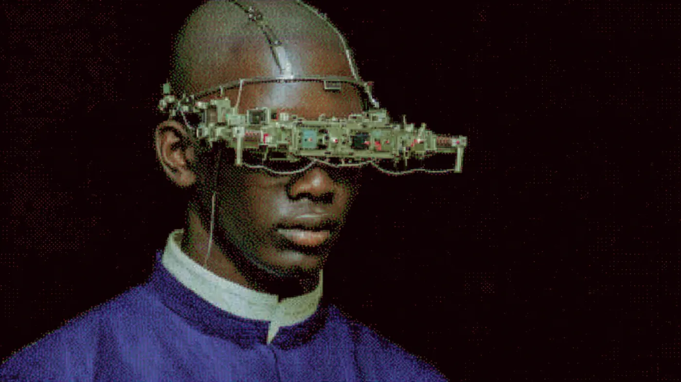 Una persona de cabeza rapada lleva un visor hecho de piezas metálicas y cables que le cubre la frente y los ojos, con cuello blanco alto y chaqueta morada, sobre un fondo oscuro.
