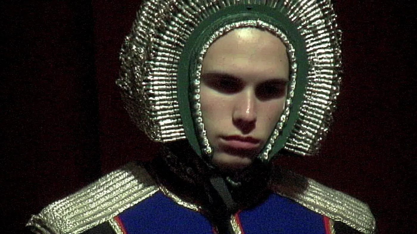 Retrato de una persona con un tocado ornamentado de cuentas plateadas y bordes verdes, vistiendo una prenda con motivos azules y plateados, sobre fondo oscuro rojizo.