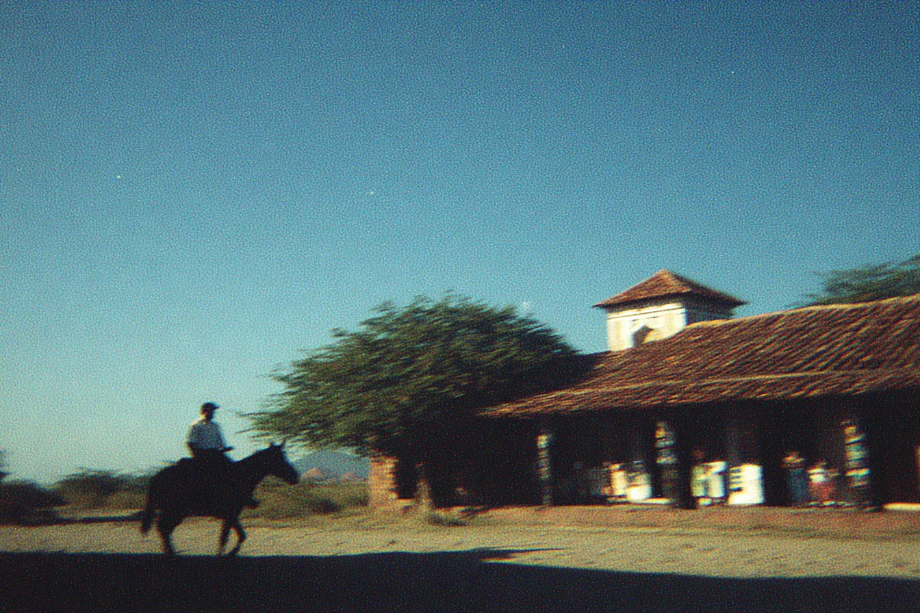 Una persona monta a caballo y pasa frente a una construcción rural de techo de paja con una pequeña torre, rodeada de árboles, bajo un cielo azul despejado durante el día.