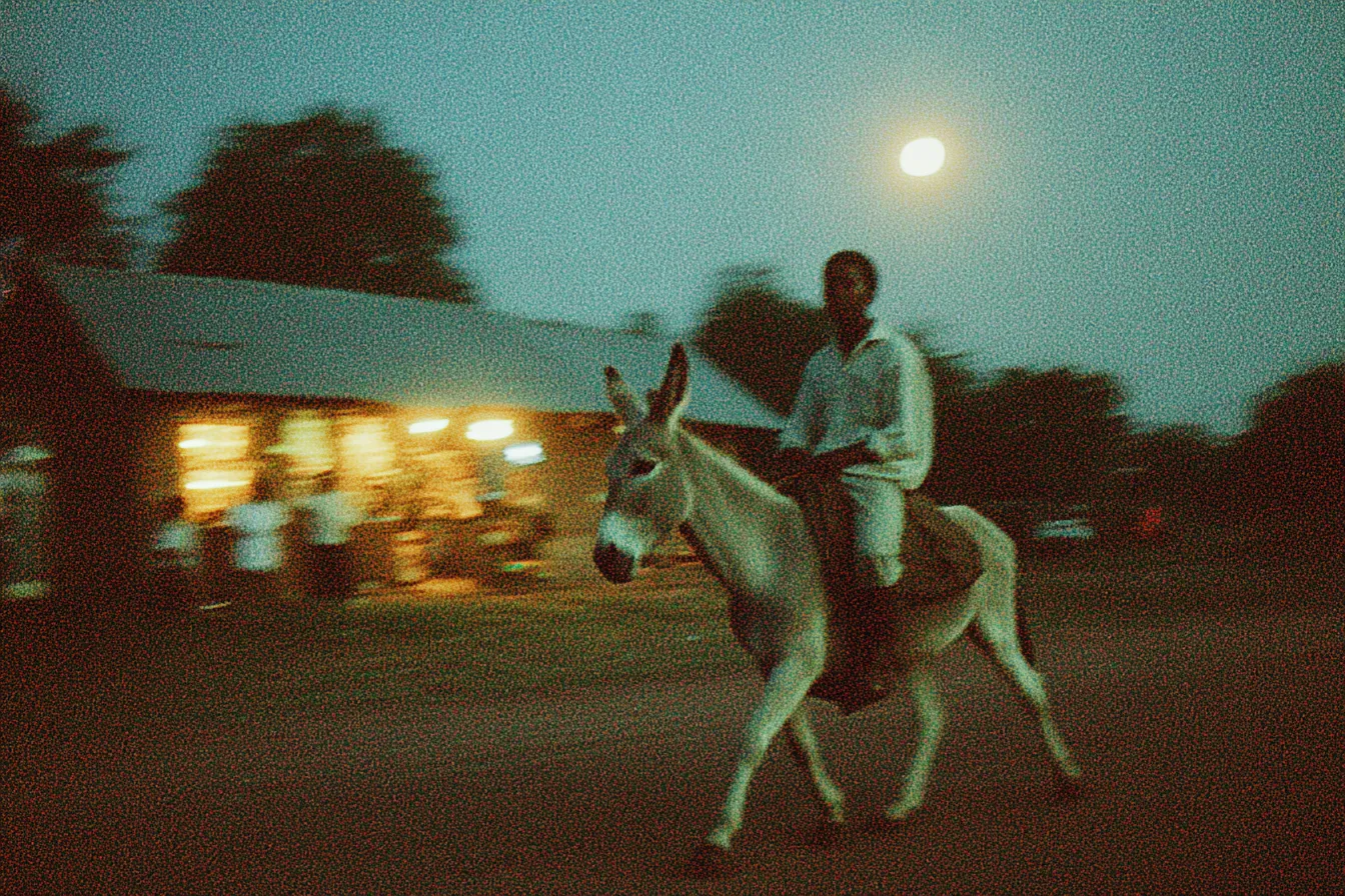 Una persona monta un burro de noche frente a una casa con las luces encendidas, bajo una luna llena, con efecto de movimiento en la imagen.
