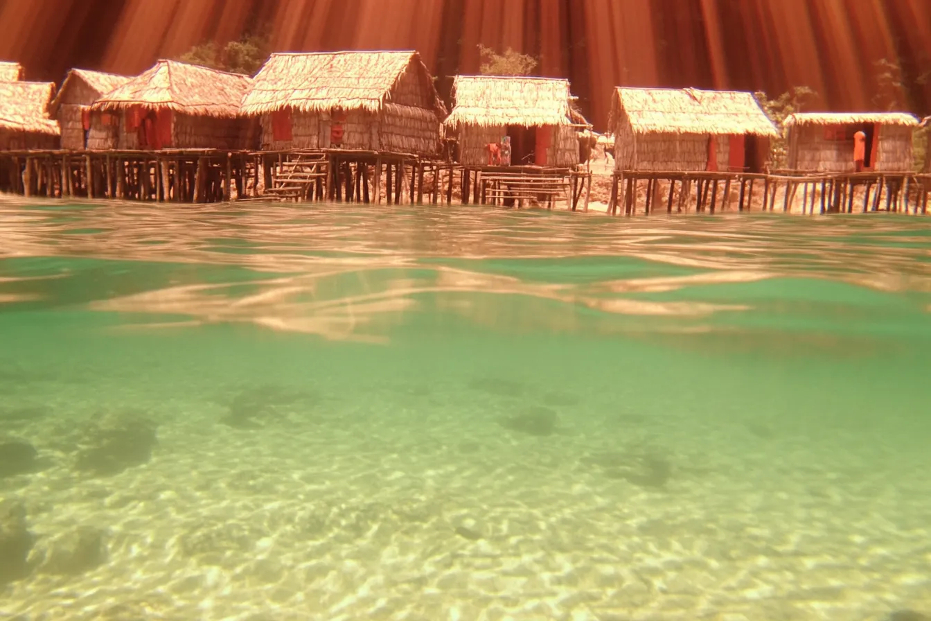 Fila de casas con techo de paja sobre pilotes en tonos cálidos anaranjados, con puertas rojas, rayos de luz cayendo desde el cielo y una imagen dividida que muestra su reflejo bajo el agua.