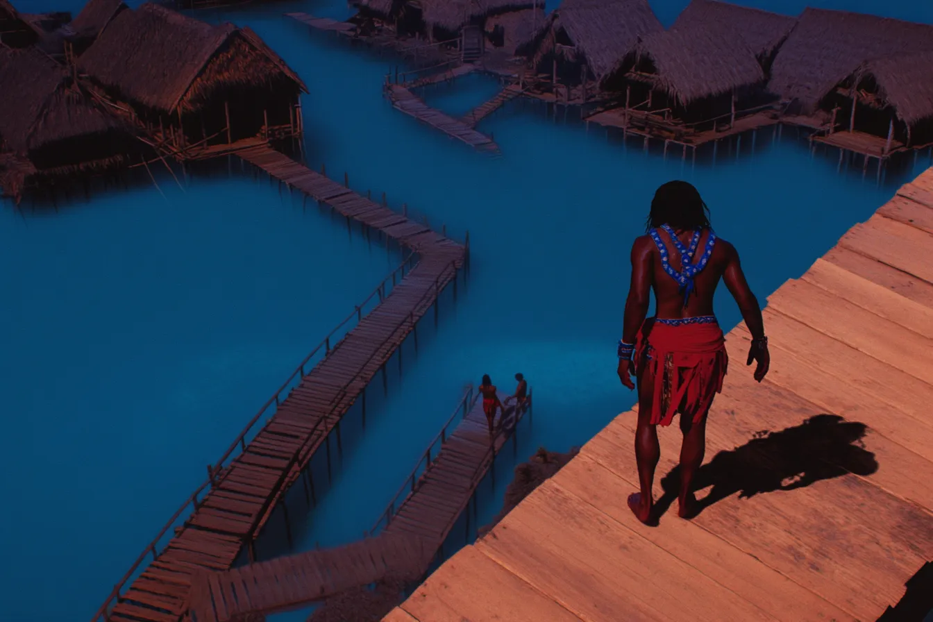 Vista elevada de una persona con collar de cuentas y falda roja caminando por un largo muelle de madera hacia un poblado de casas con techo de paja sobre el agua, en tonos azules y violetas de atardecer, con su sombra proyectada sobre las tablas.