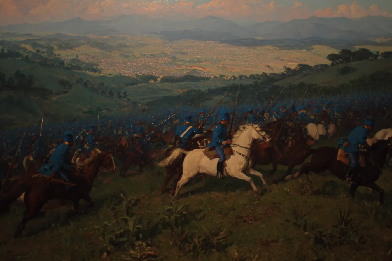 Pintura de una batalla de caballería en una colina al atardecer, con jinetes de uniforme azul en primer plano, humo disperso, y un pueblo pequeño visible en el valle iluminado por luz dorada.