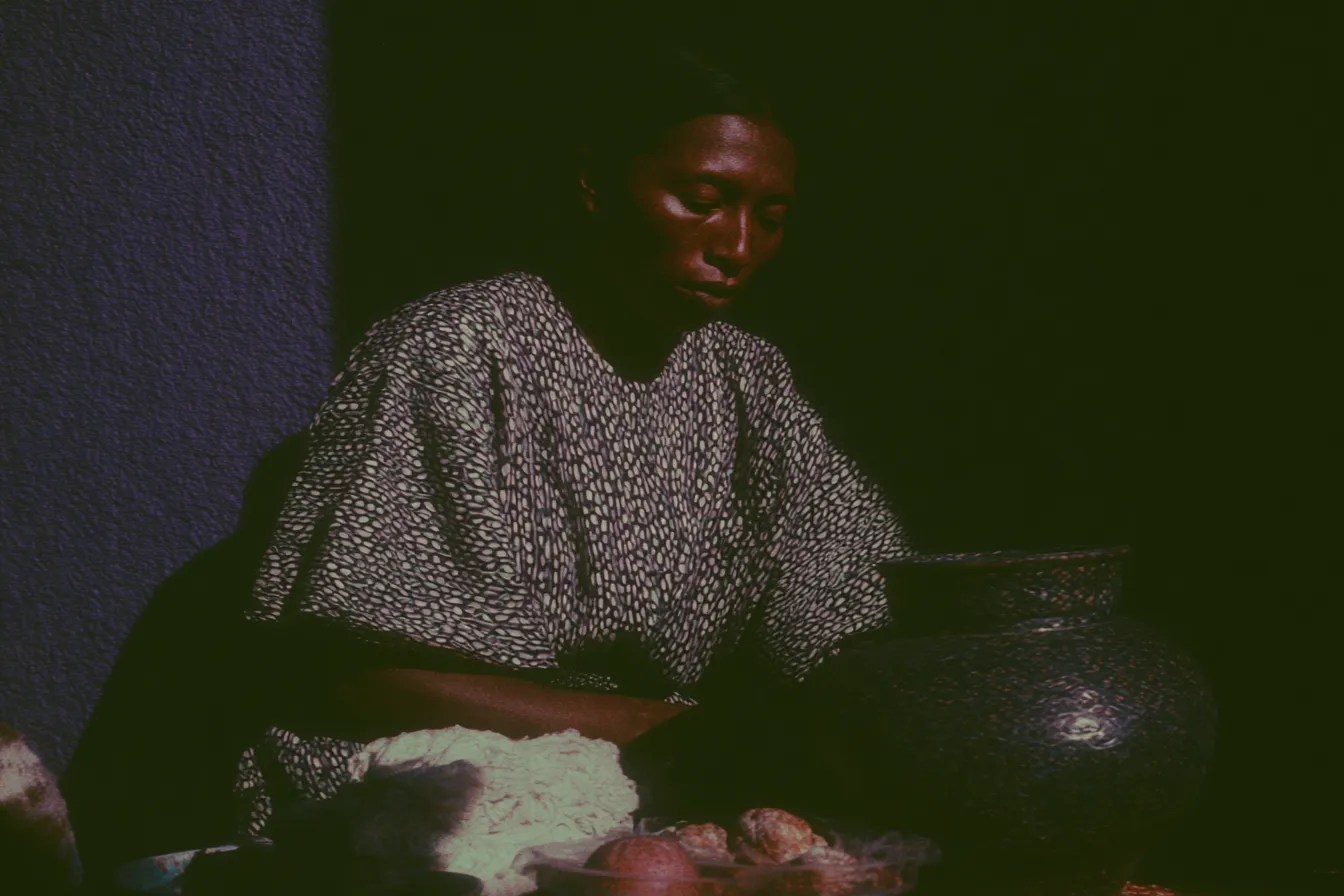 Fotografía oscura de una mujer con vestido estampado sentada junto a una gran olla de barro negro, con alimentos dispuestos cerca, en un ambiente de escasa iluminación.