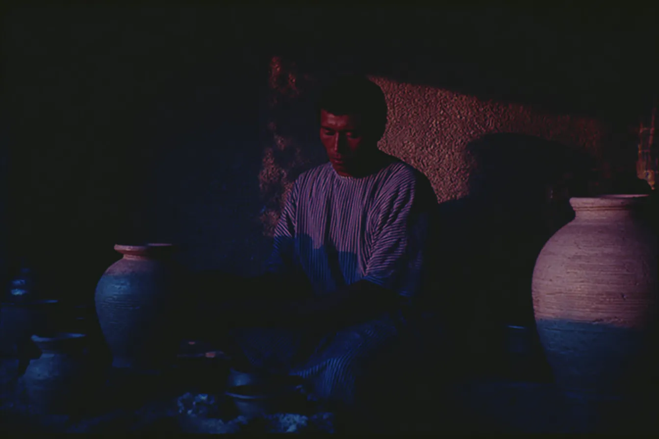 Fotografía oscura de un hombre sentado en el suelo entre dos grandes vasijas de barro, iluminado por luces de tono rojo y azul que crean un ambiente de contraste dramático.