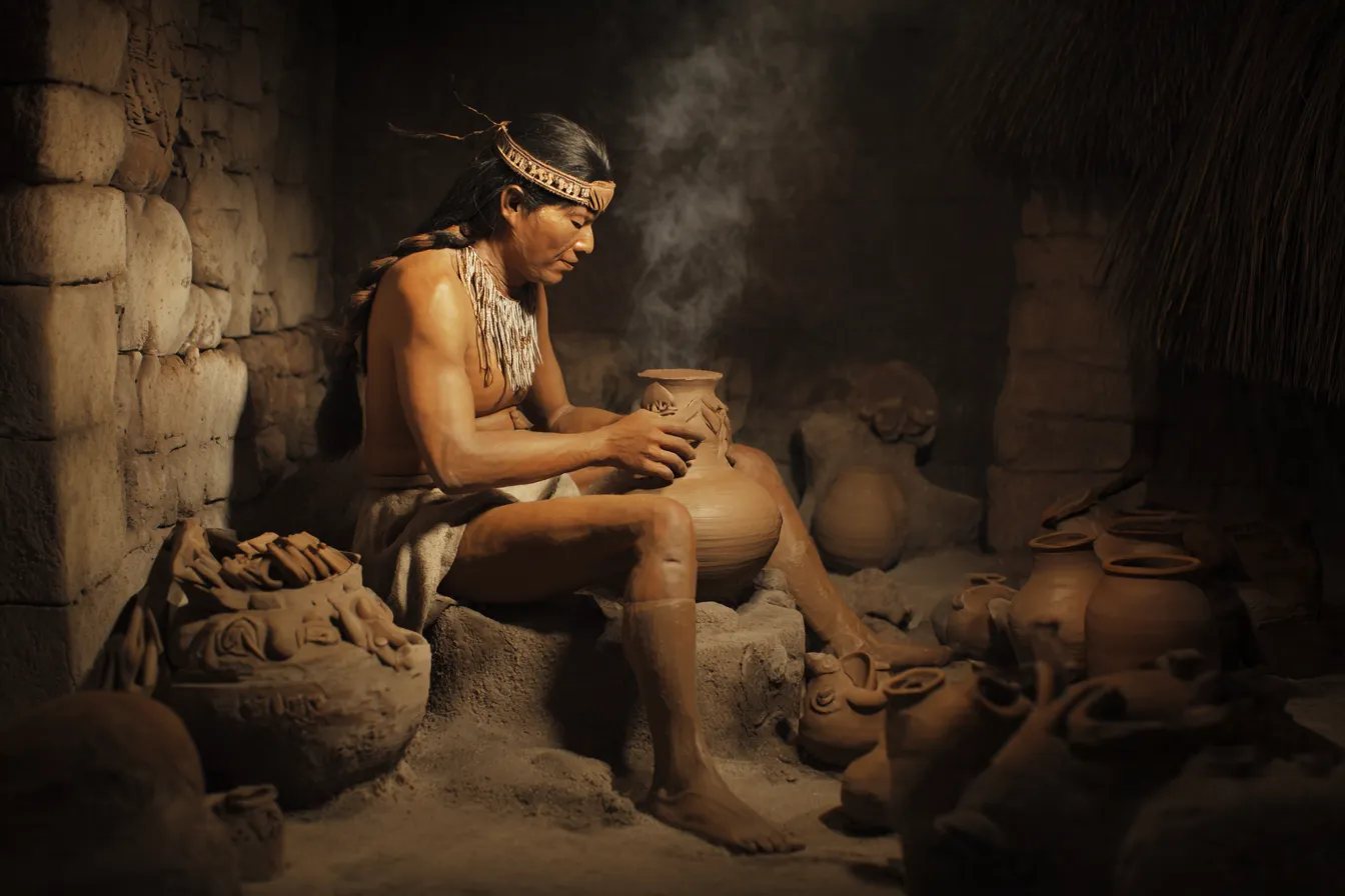 Fotografía de un hombre indígena sentado con las piernas cruzadas, moldeando una vasija de barro con las manos, con el torso descubierto, una vincha de plumas en la cabeza, humo fino elevándose del recipiente, rodeado de numerosas piezas de cerámica y una pared de piedra al fondo.
