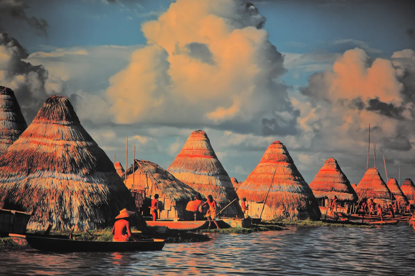 Aldea de chozas cónicas de paja construida sobre el agua, con varias personas remando en canoas vestidas de naranja y rojo, bajo un cielo de nubes doradas y anaranjadas al atardecer.