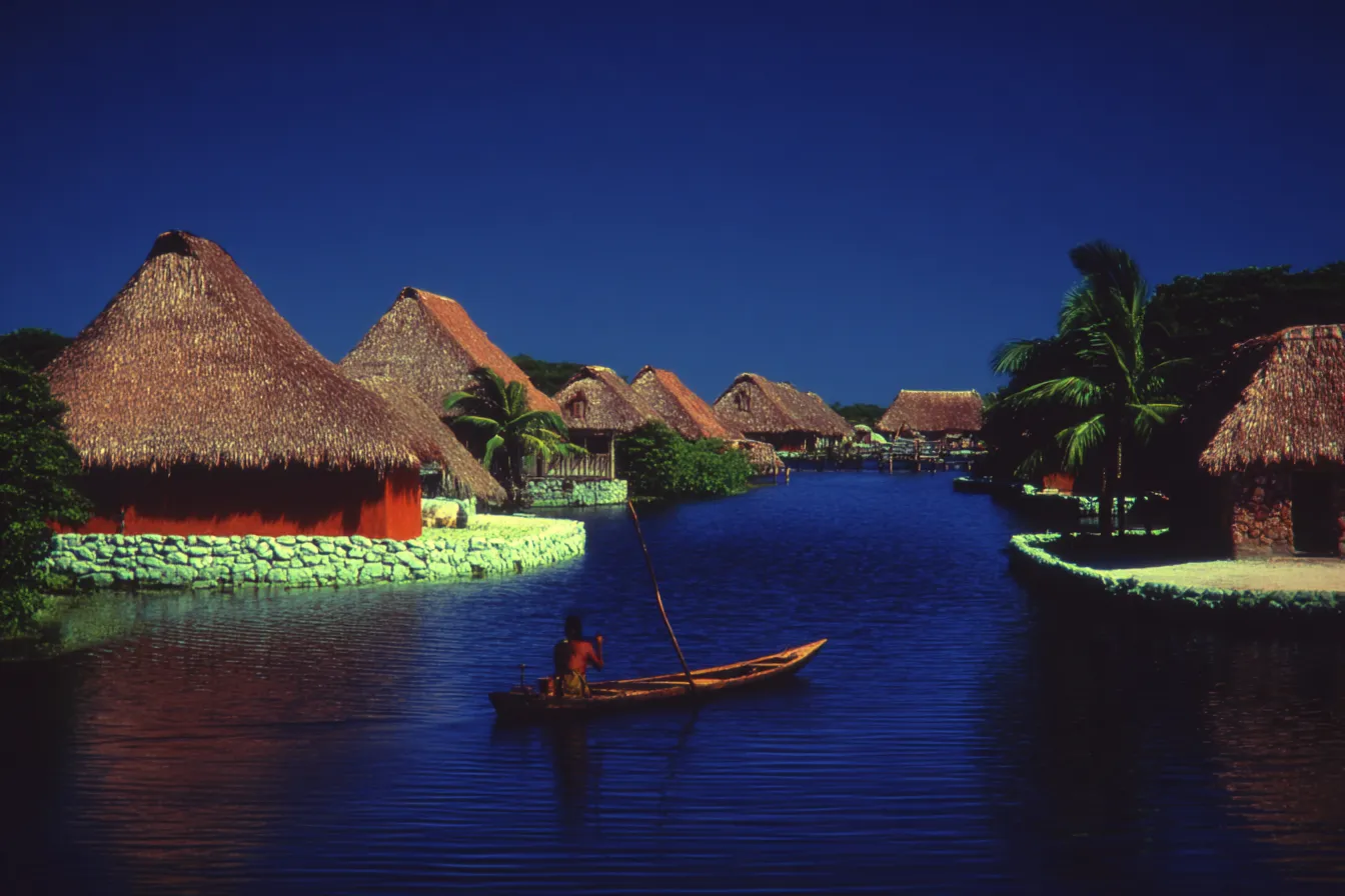 Vista nocturna de un canal entre casas de techo de paja con paredes de piedra pintadas de rojo y blanco, una persona de pie remando una canoa de madera, palmera a la derecha, cielo azul oscuro.
