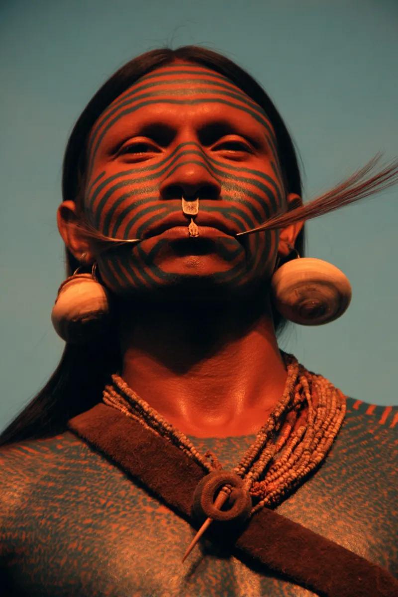 Retrato de un hombre con la cara pintada con líneas verdes onduladas, grandes discos blancos colgando de las orejas, un mechón de plumas junto a la barbilla, un collar de cuentas ajustado al cuello y una banda tejida cruzada sobre el pecho, sobre fondo azul verdoso.
