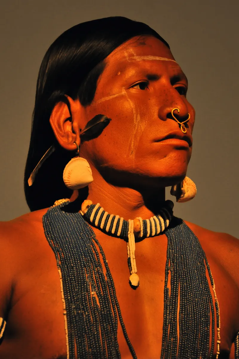 Retrato de un hombre de cabello negro liso con una raya blanca pintada verticalmente en el centro del rostro, un gran aro dorado en el tabique nasal, aretes de disco con una pluma y varios collares de cuentas oscuras, sobre fondo gris oscuro.