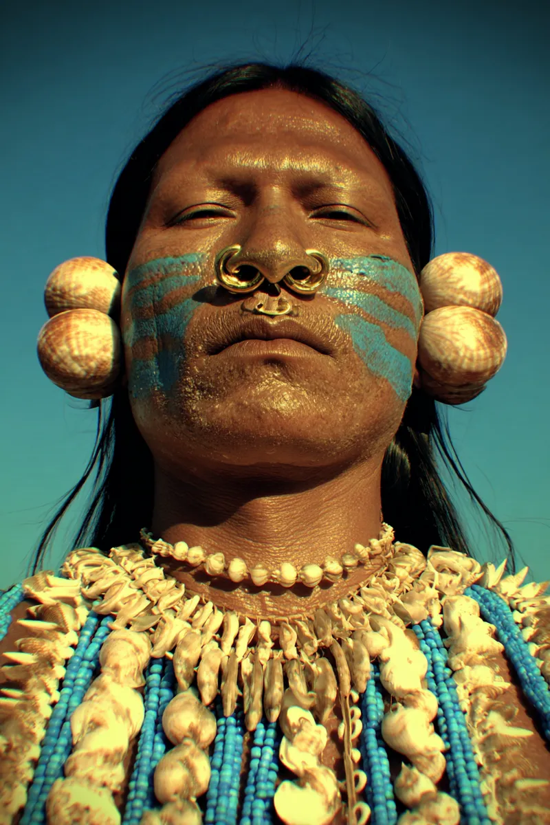 Retrato de un hombre con los ojos cerrados, rayas de pintura turquesa en las mejillas, un doble aro dorado en la nariz, grandes discos de concha en las orejas y un collar de conchas y cuentas, sobre fondo de cielo azul, con la piel iluminada en tono dorado.