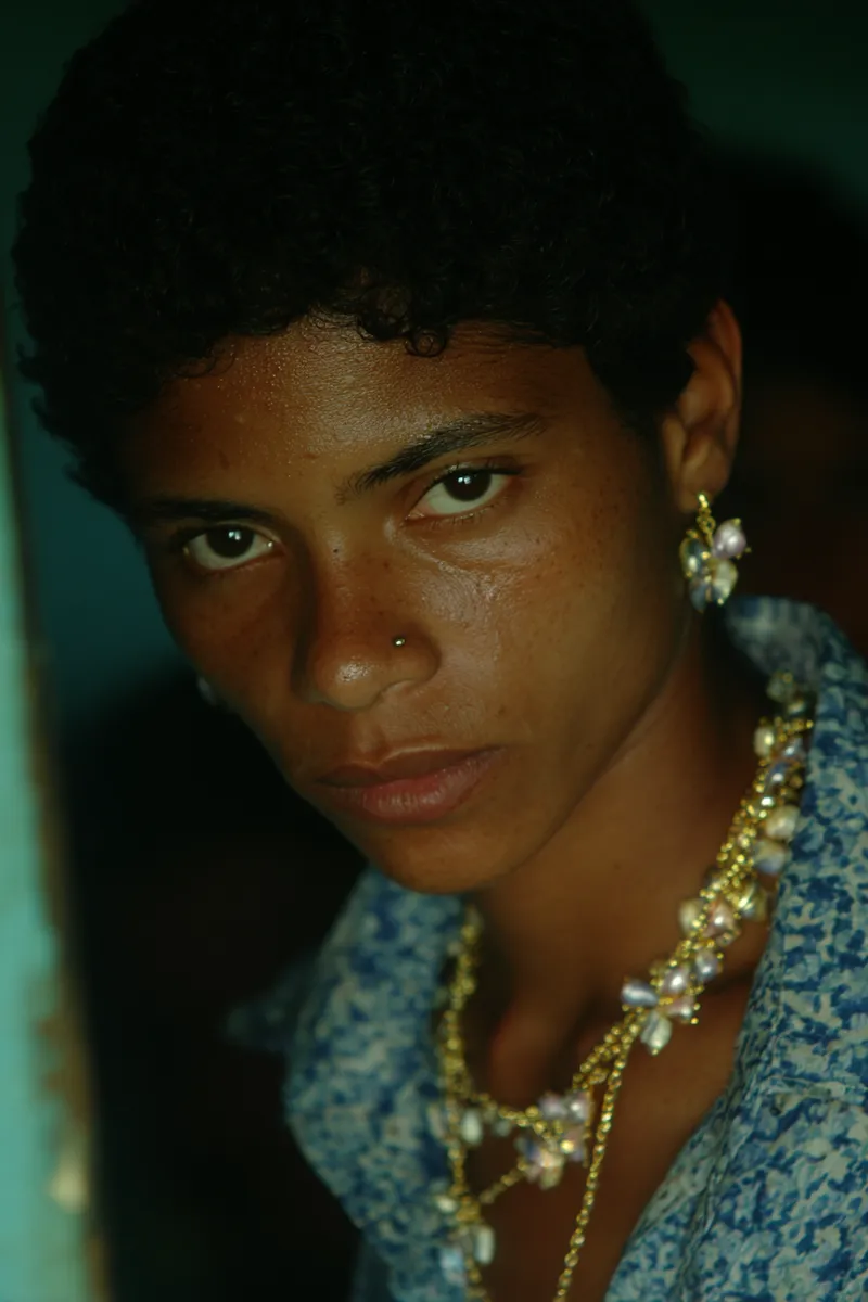 Retrato de una joven de cabello oscuro y rizado, con aretes de perla y un collar dorado, vestida con una blusa azul de flores, mirando directamente a la cámara con el rostro iluminado de un lado por luz lateral junto a un marco de puerta azul.