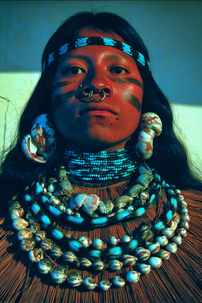 Retrato de una mujer con una diadema de cuentas, la cara pintada con rayas naranjas y negras, un aro en el tabique nasal, grandes aretes de concha y varios collares de cuentas azules y conchas, frente a un fondo de esteras tejidas.