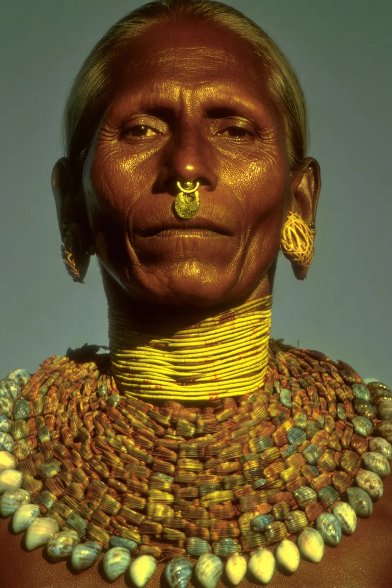 Retrato de una mujer mayor con el rostro marcado por arrugas, un aro dorado en la nariz, aretes dorados de aro y el cuello cubierto de anillos apretados de cuentas amarillas, además de collares de cuentas y conchas de varios colores.