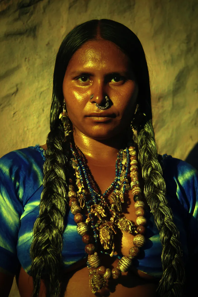 Retrato de una mujer con dos trenzas largas de cabello, vestida con una tela a rayas azules y verdes, un aro dorado en la nariz y varios collares de cuentas con un colgante dorado, iluminada con luz cálida contra una pared de textura rugosa.