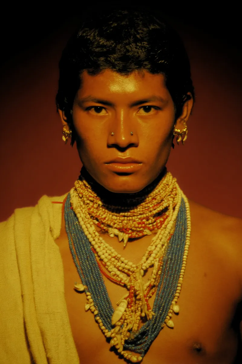 Retrato de un joven de cabello rizado con pequeños aretes dorados de aro, un aro en la nariz y varios collares de cuentas azules, rojas y blancas, con una tela clara envuelta sobre un hombro, contra un fondo rojo oscuro.