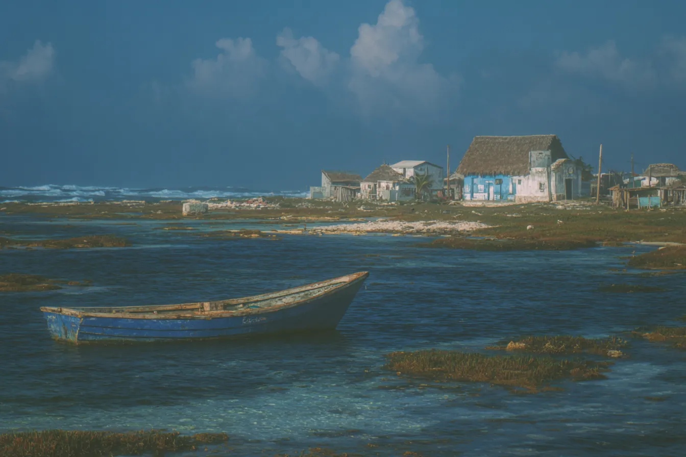 Bote de madera azul varado sobre un arrecife de coral en aguas turquesas poco profundas, con casas de techo de paja en la orilla bajo un cielo nublado.