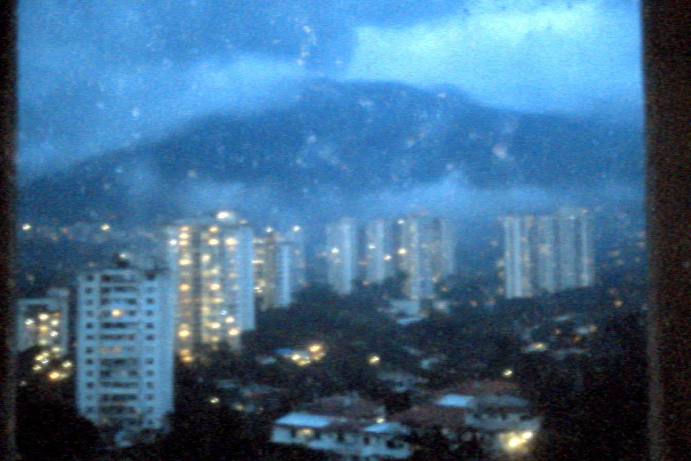Vista nocturna de una ciudad con edificios iluminados y una montaña cubierta de nubes al fondo, fotografiada a través de un vidrio salpicado de gotas de lluvia.