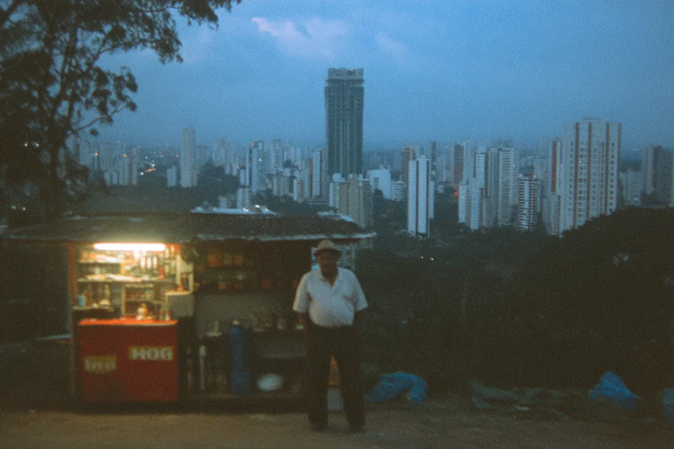 Hombre de pie junto a un quiosco de comida iluminado, con una nevera roja al frente y una hilera de edificios altos iluminados detrás al anochecer.