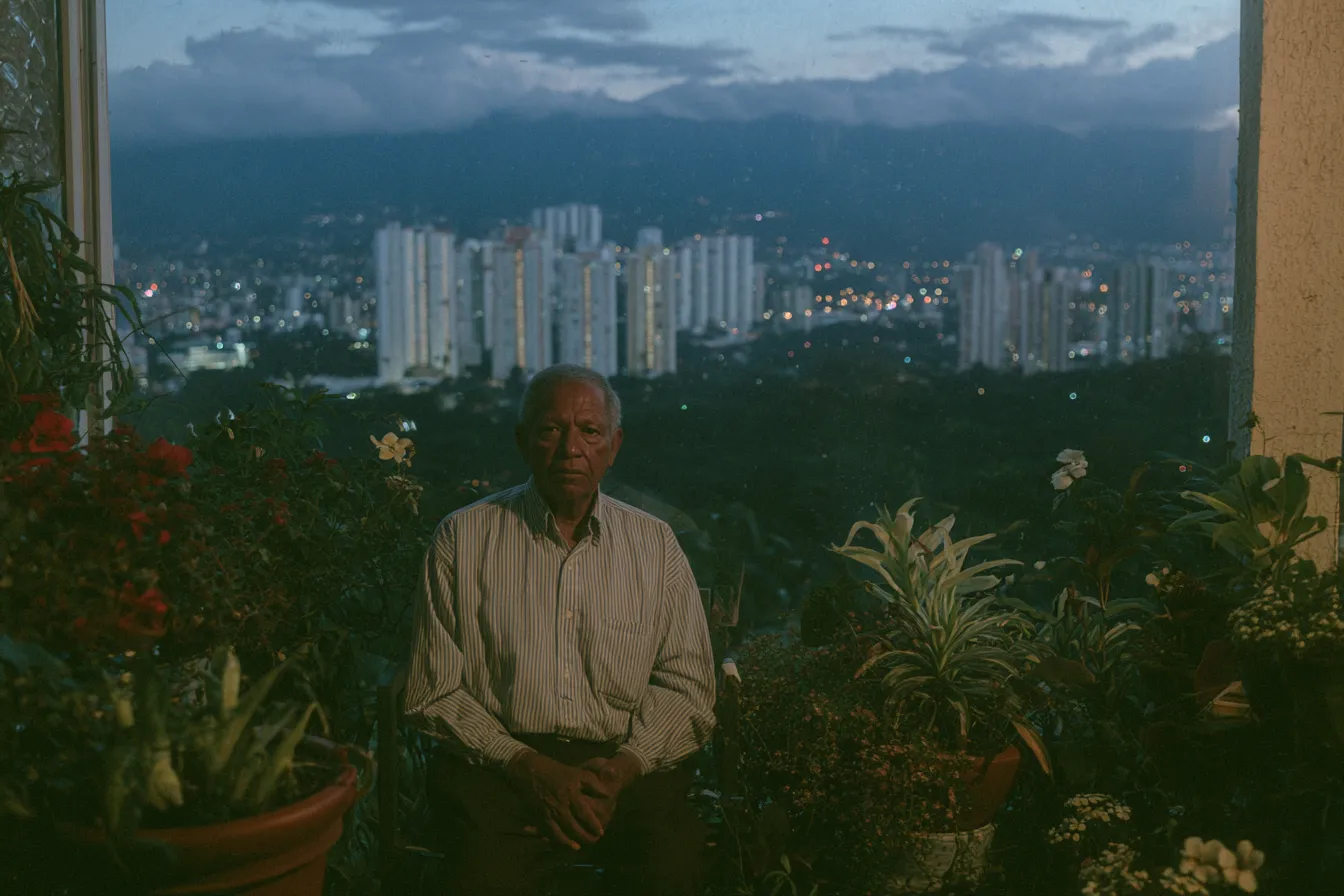 Hombre mayor de pie en un balcón rodeado de macetas con flores, con vista a una ciudad iluminada y montañas al fondo durante el atardecer.