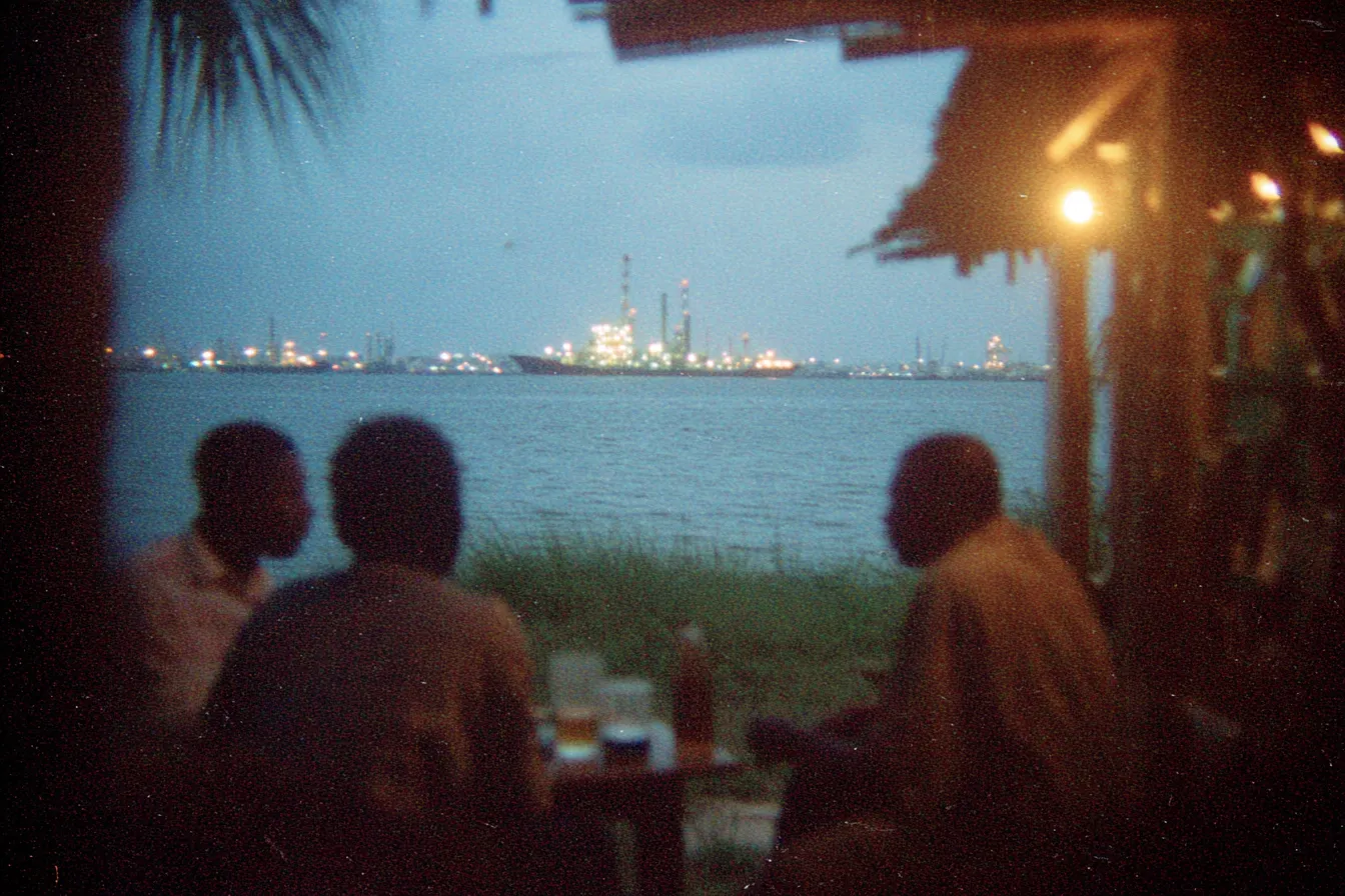 Tres hombres sentados de espaldas bajo un techo de palma al anochecer, mirando hacia una refinería industrial iluminada al otro lado del agua.