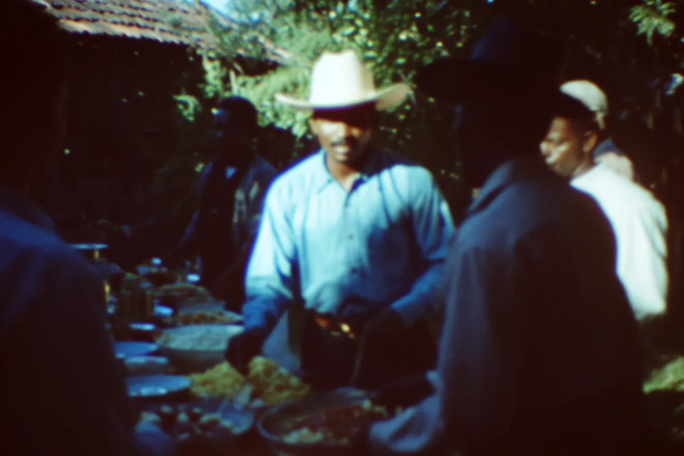 Hombre con sombrero blanco y camisa azul sonríe mientras sirve comida desde bandejas grandes, rodeado de otras personas bajo un techo de palma.