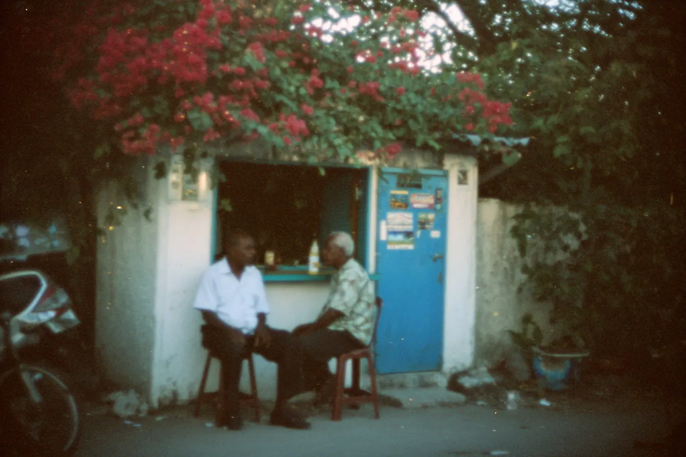 Dos hombres conversan sentados en sillas frente a una puerta azul, bajo una buganvilia con flores rojas, junto a una motocicleta estacionada.