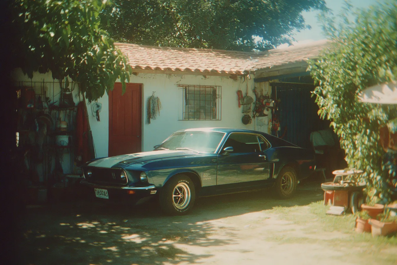 Automóvil deportivo verde oscuro estacionado frente a una casa blanca de techo de tejas con puerta roja, bajo la sombra de un árbol en un día soleado.