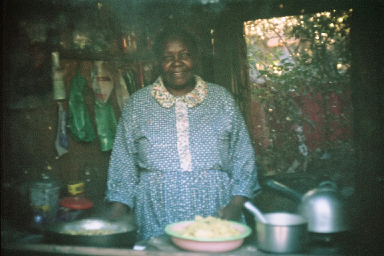 Mujer mayor sonriente con vestido floral y cuello de encaje, de pie frente a una mesa de cocina con un plato de arroz y varias ollas.