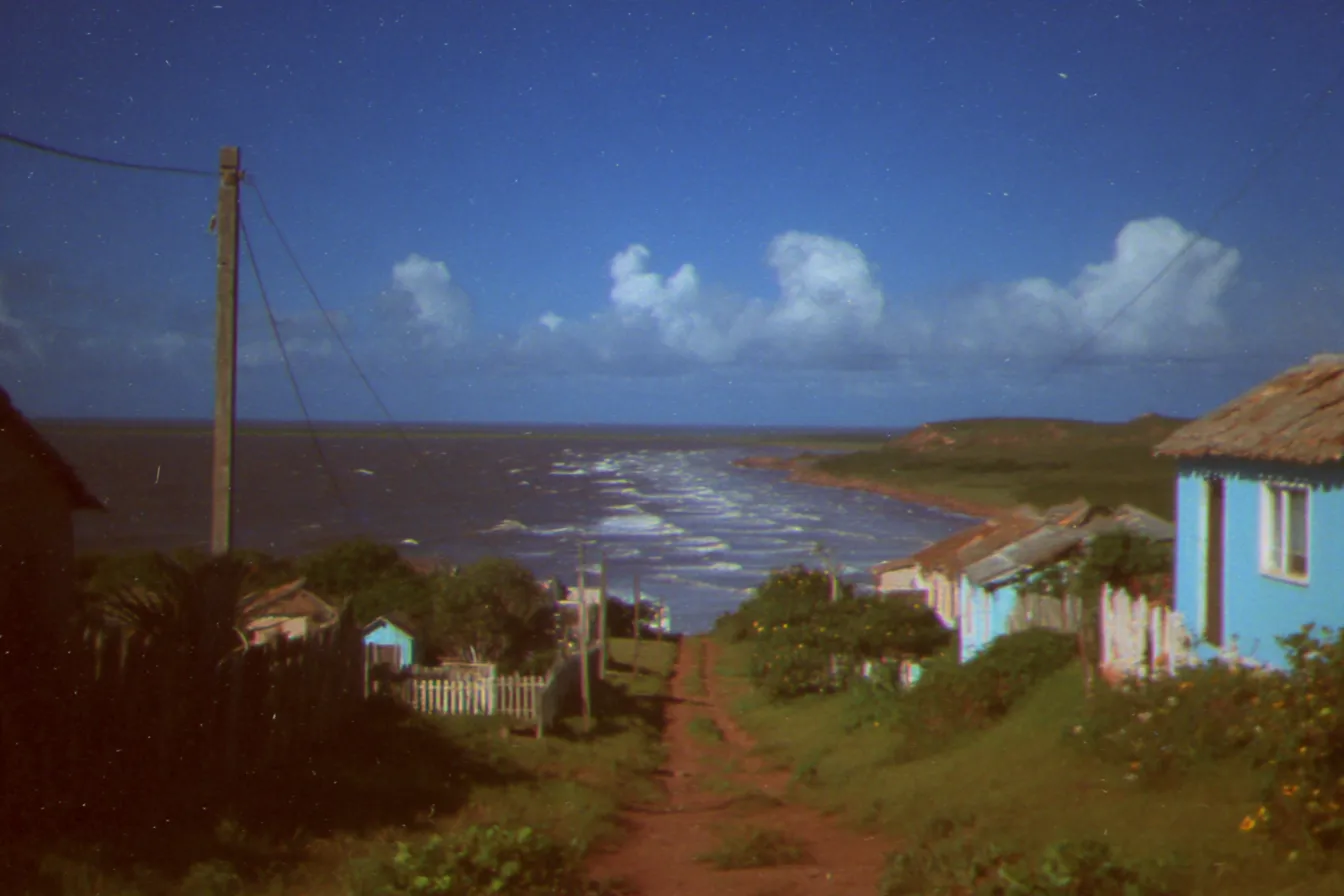 Camino de tierra roja que baja entre casas de techo de paja hasta una bahía de noche, con una casa azul y una cerca blanca a un lado, bajo un cielo estrellado.