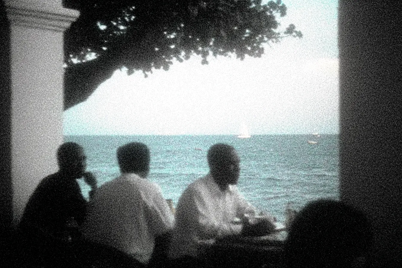 Tres hombres con camisas blancas conversan sentados a una mesa bajo un árbol grande, en una terraza con columnas, frente al mar donde se ven veleros a lo lejos.