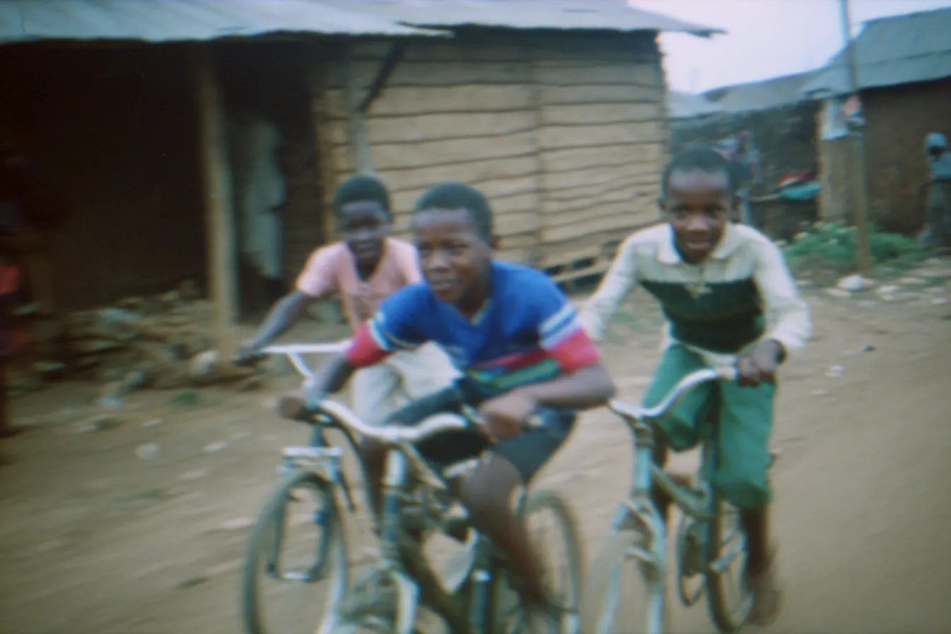 Tres niños montan bicicletas por un camino de tierra frente a casas de tablones de madera con techo de zinc; el que va adelante viste camiseta azul, roja y blanca.