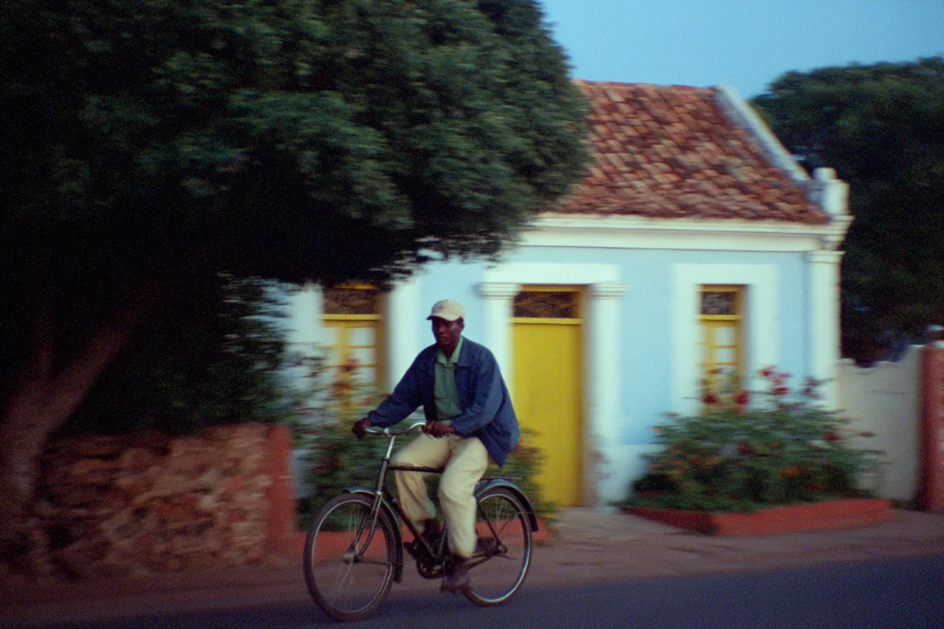 Hombre con gorra monta una bicicleta frente a una casa colonial de paredes claras, puerta amarilla y techo de tejas, al atardecer, con flores rojas junto a la reja.