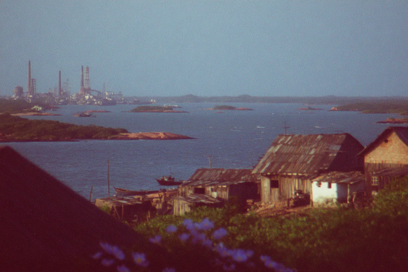 Vista de un golfo con una refinería industrial y sus chimeneas al fondo, botes pequeños en el agua, y en primer plano casas rústicas de techo de paja y flores moradas.