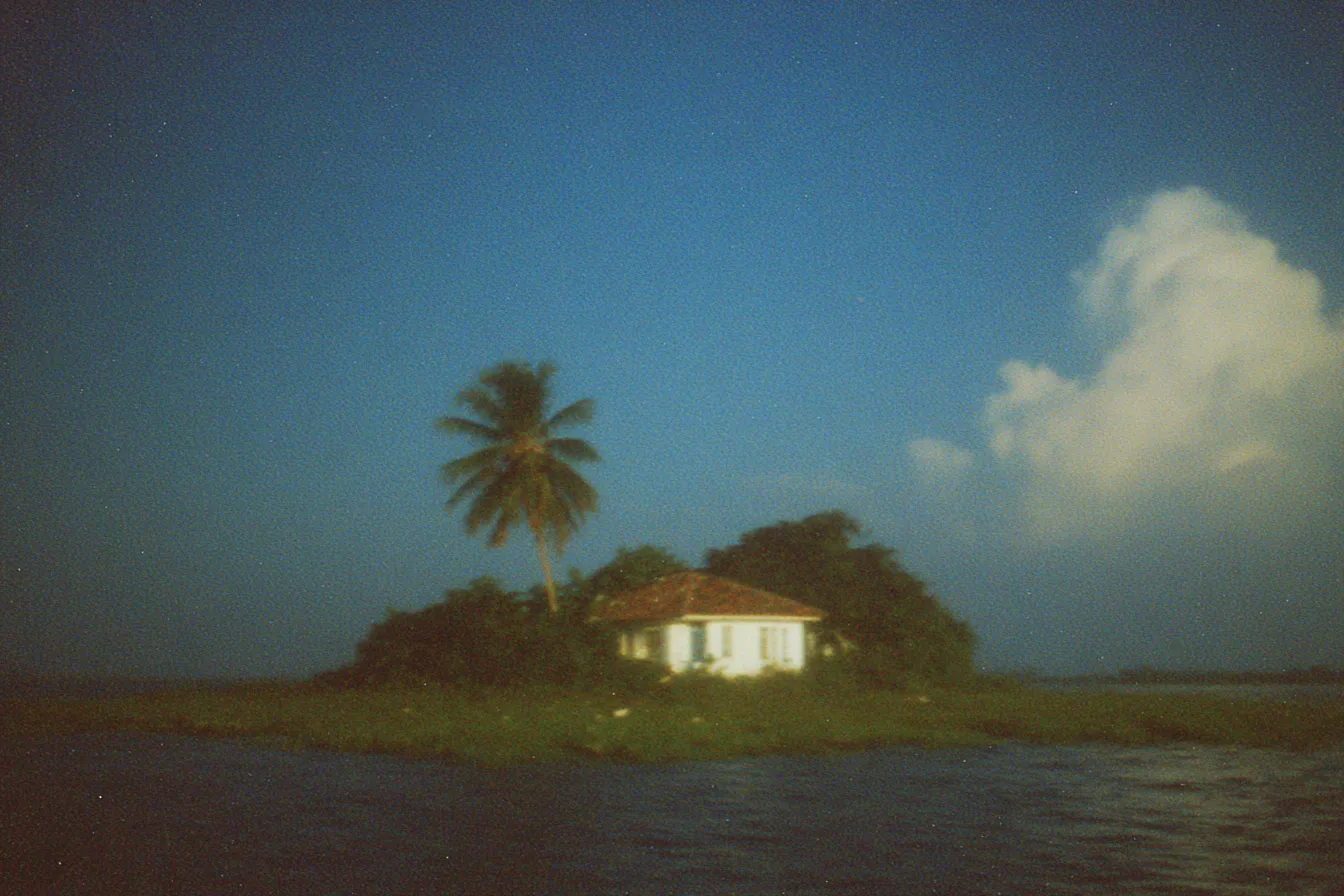 Casa blanca pequeña y una palmera sobre un islote rodeado de agua, bajo un cielo azul con una nube grande.