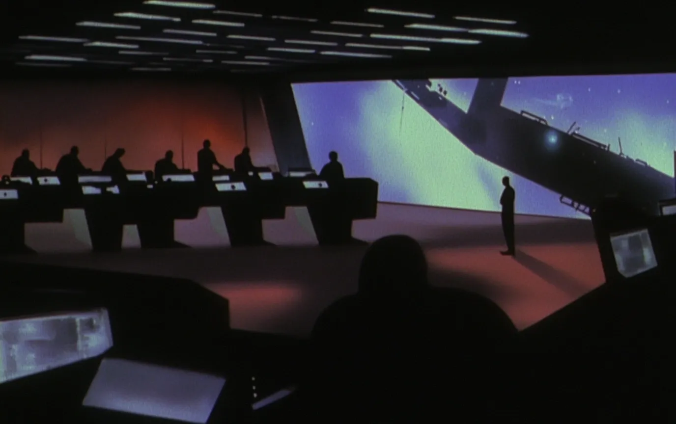 Sala de control con una fila de personas sentadas ante consolas iluminadas, una persona de pie mirando una pantalla gigante con una forma blanca y azul sobre fondo de estrellas, piso con tono anaranjado.